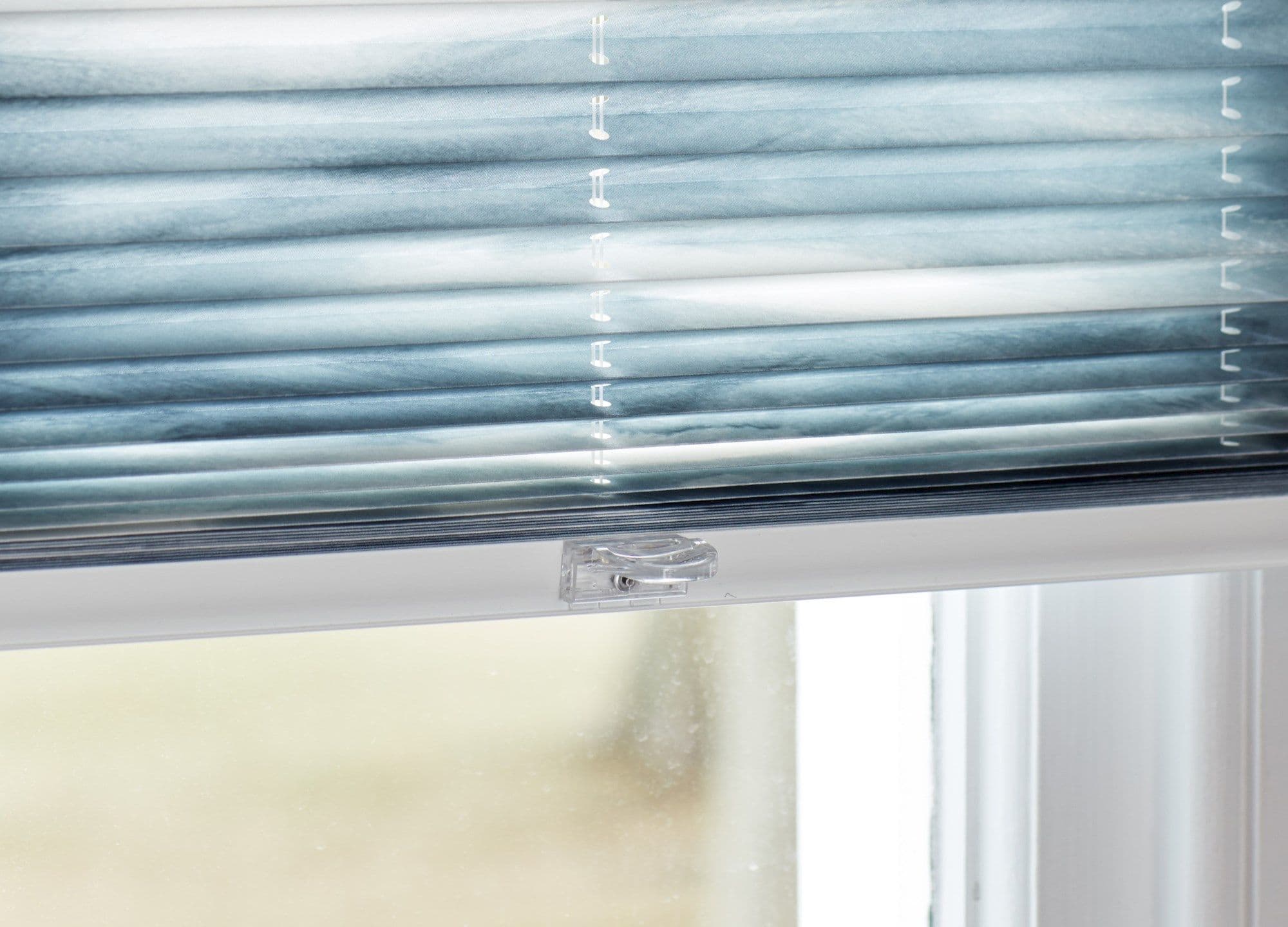Blue pleated cellular blind, lowered with visible bottom rail and central clear plastic latch â€” filtering soft daylight â€” interior window with a blurred pale outdoor view.