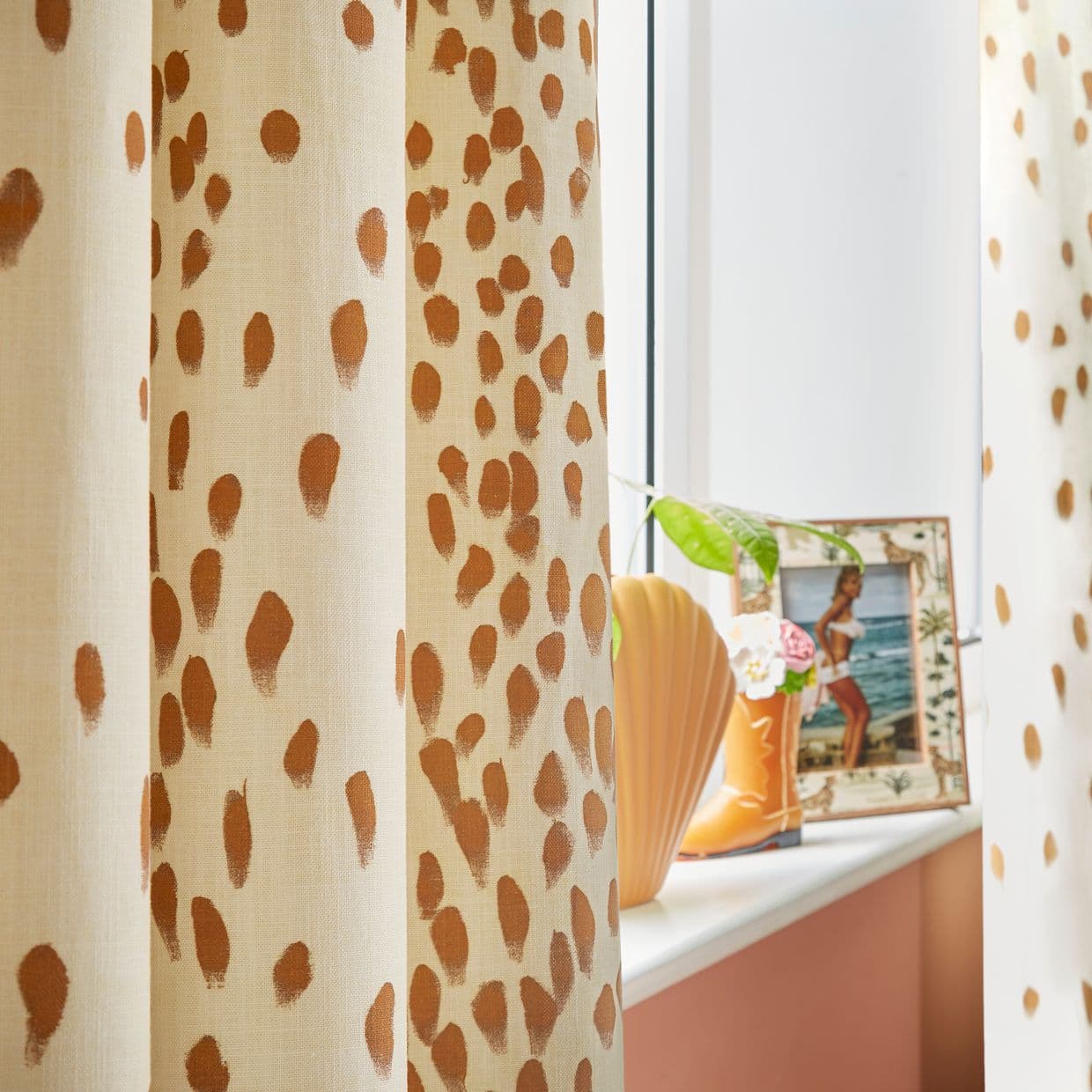 Cream curtains with terracotta spots, hanging partly drawn and filtering daylight; on the windowsill a shell-shaped vase, boot planter with flowers and a framed beach photo in a warm-toned room.