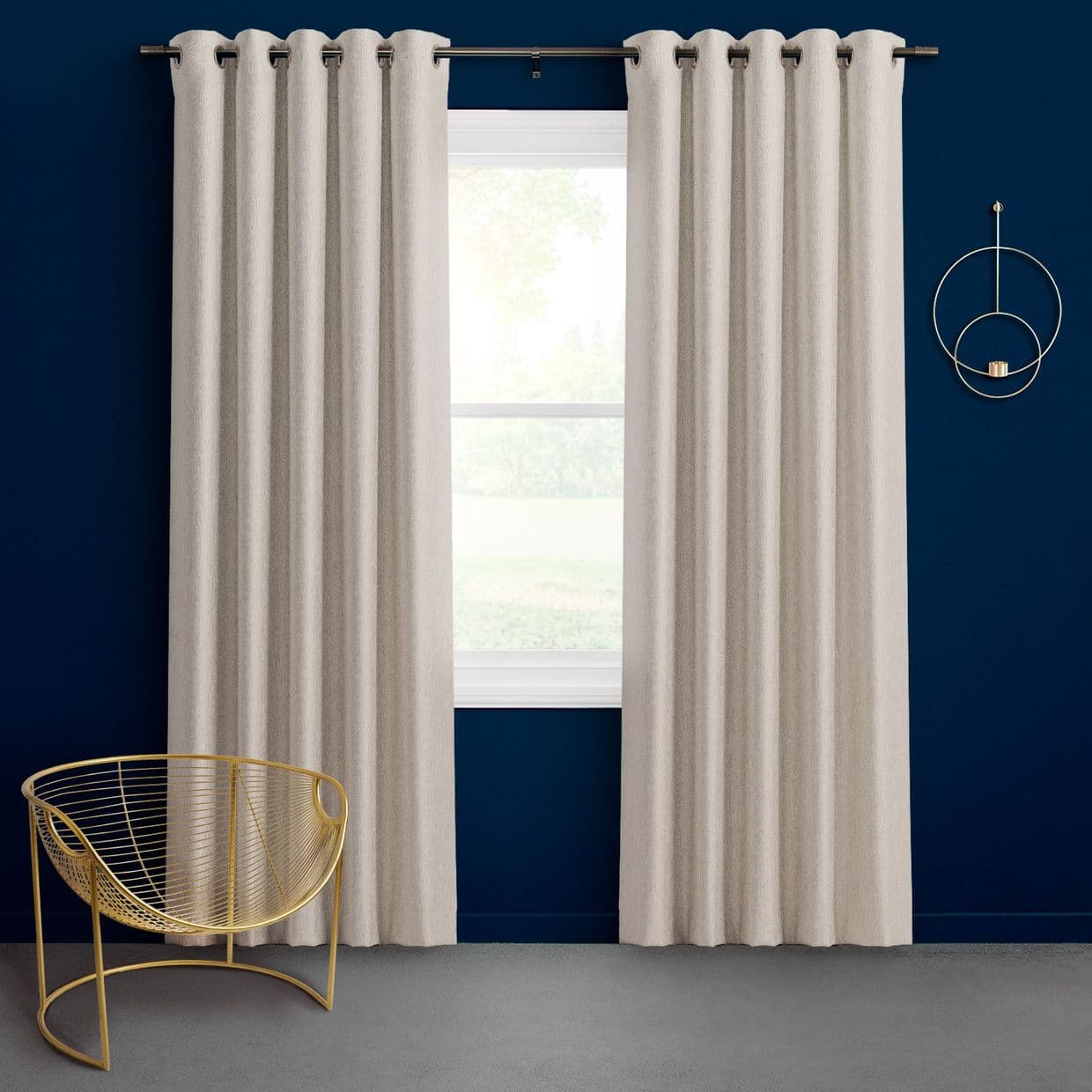 Cream grommet-top curtains, hung on a metal rod and parted to reveal a central window, filtering soft daylight; set against a deep navy wall with a gold wire chair.