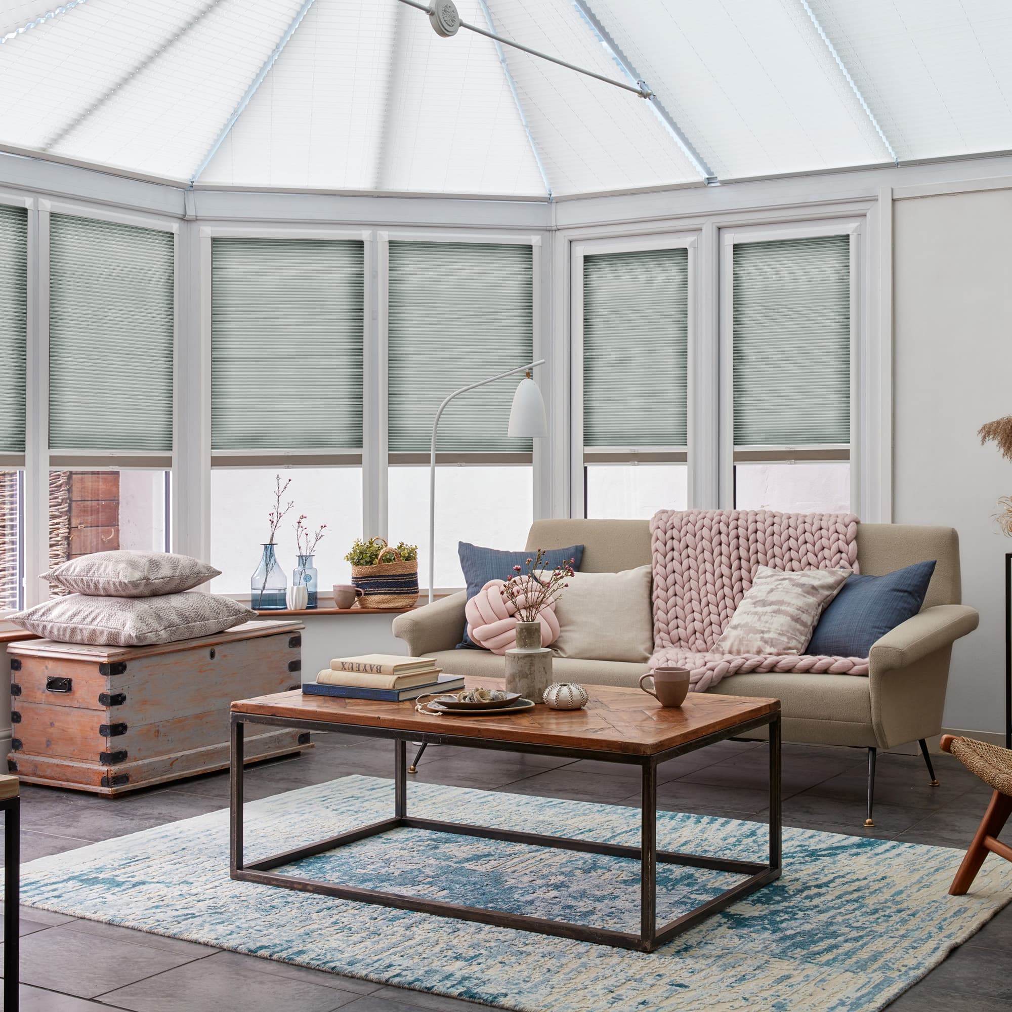 Pleated cellular blinds covering tall conservatory windows, lowered to mid-height and softly filtering daylight; sunroom with beige sofa, chunky pink knitted throw, wooden coffee table, decorative vases on the sill.