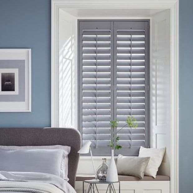 Grey plantation-style shutters (slatted panels) closed, filtering light and casting striped shadows; set in a white recessed window with a cushioned seat; pale-blue bedroom with bed, small table, vase.