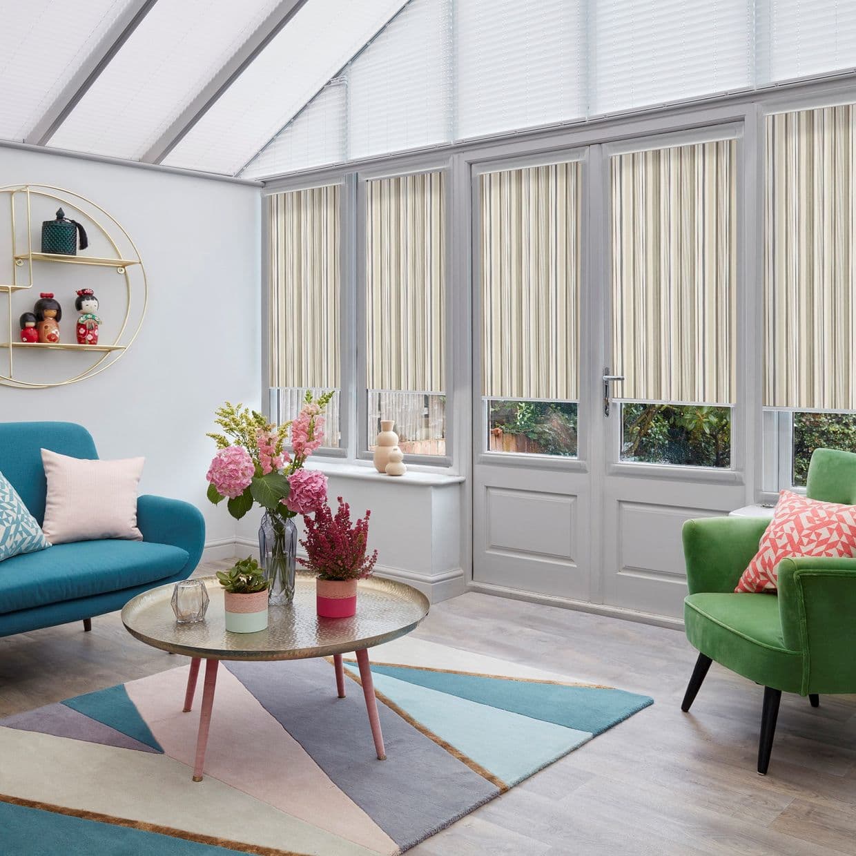 Striped beige vertical blinds on tall conservatory windows and French doors, lowered to cover upper panes and filter daylight, in a bright sitting room with glass roof, sofas and rug.