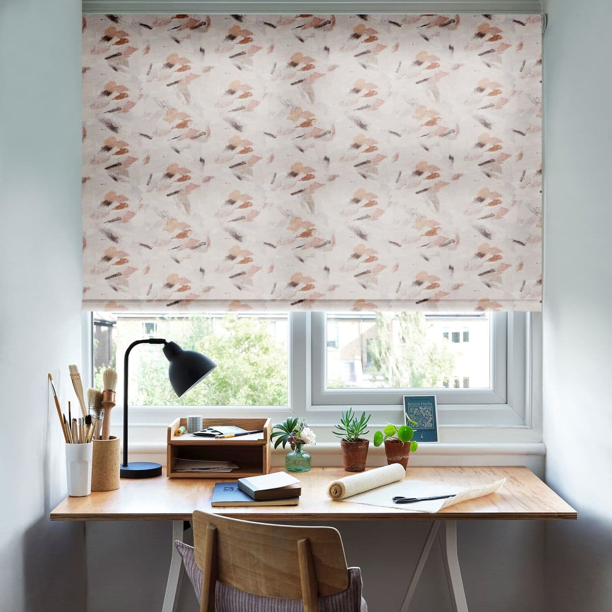 A patterned roller blind (soft pinkâ€‘beige brushstrokes) is lowered over the window, diffusing daylight; below, a wooden desk holds a lamp, art brushes, notebooks and potted plants, with trees outside.