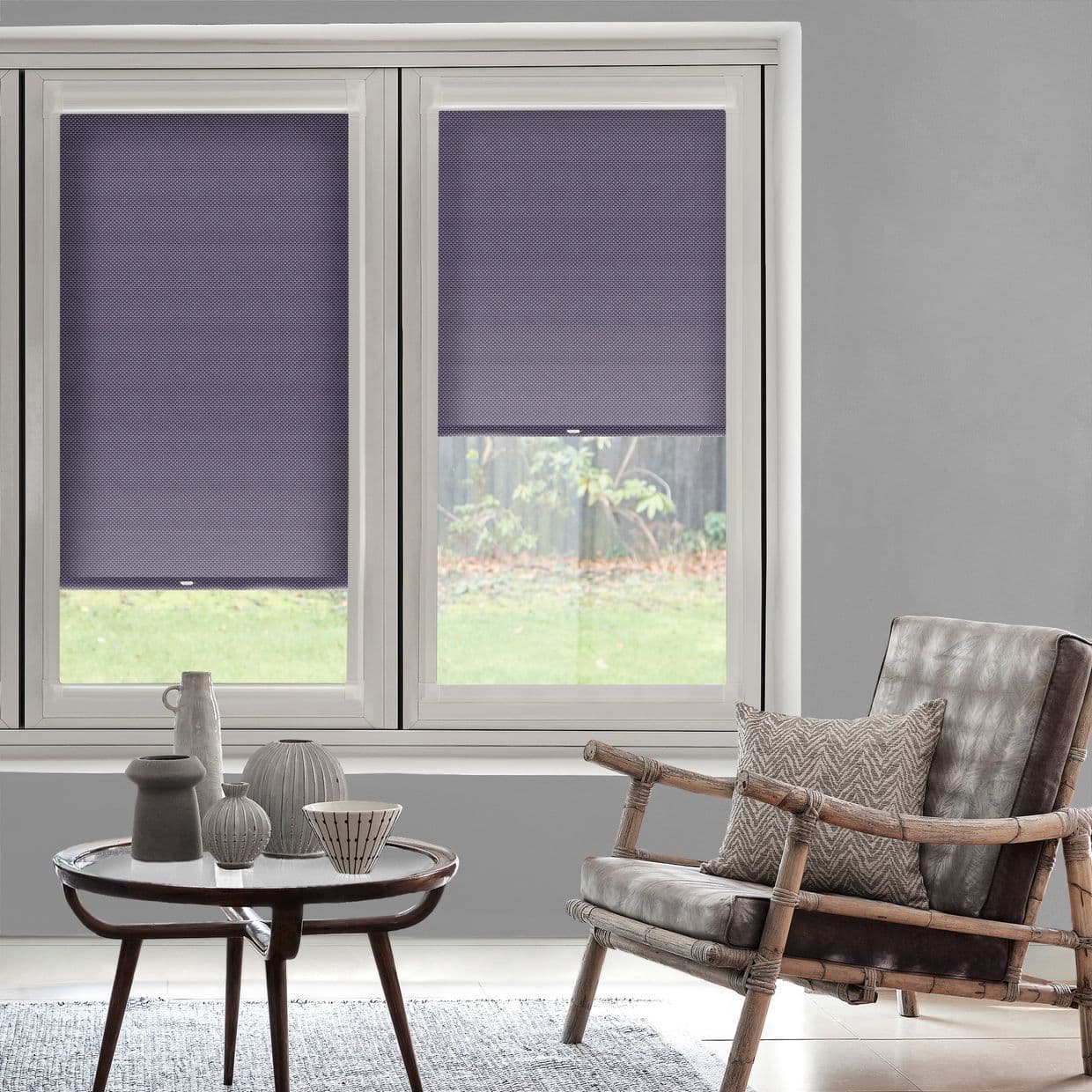Purple honeycomb cellular blinds on two adjacent window panesâ€”left fully lowered, right halfway raisedâ€”filter daylight and obscure a backyard view in a softly lit gray living room with rattan armchair.