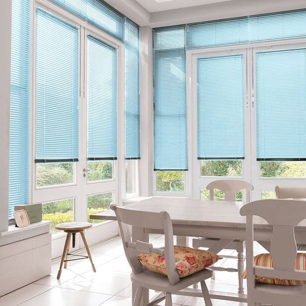 Light-blue horizontal venetian blinds covering tall floor-to-ceiling windows, largely lowered and filtering soft daylight; bright white dining sunroom with wooden table, chairs and garden view.