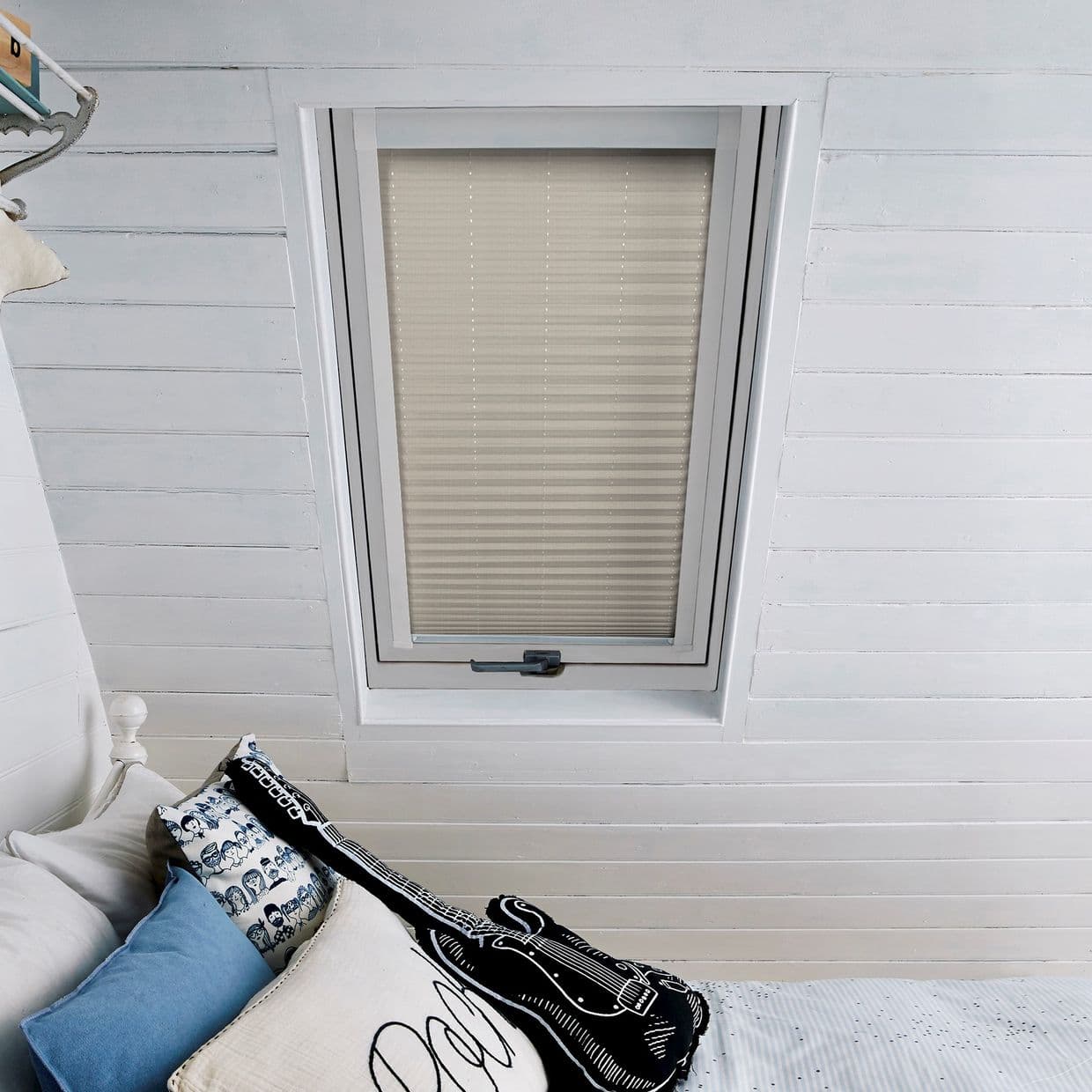 Beige pleated blind in a rectangular skylight, fully closed with horizontal folds and perforations, filtering light over a white-painted attic ceiling and a bed with pillows and a guitar cushion.