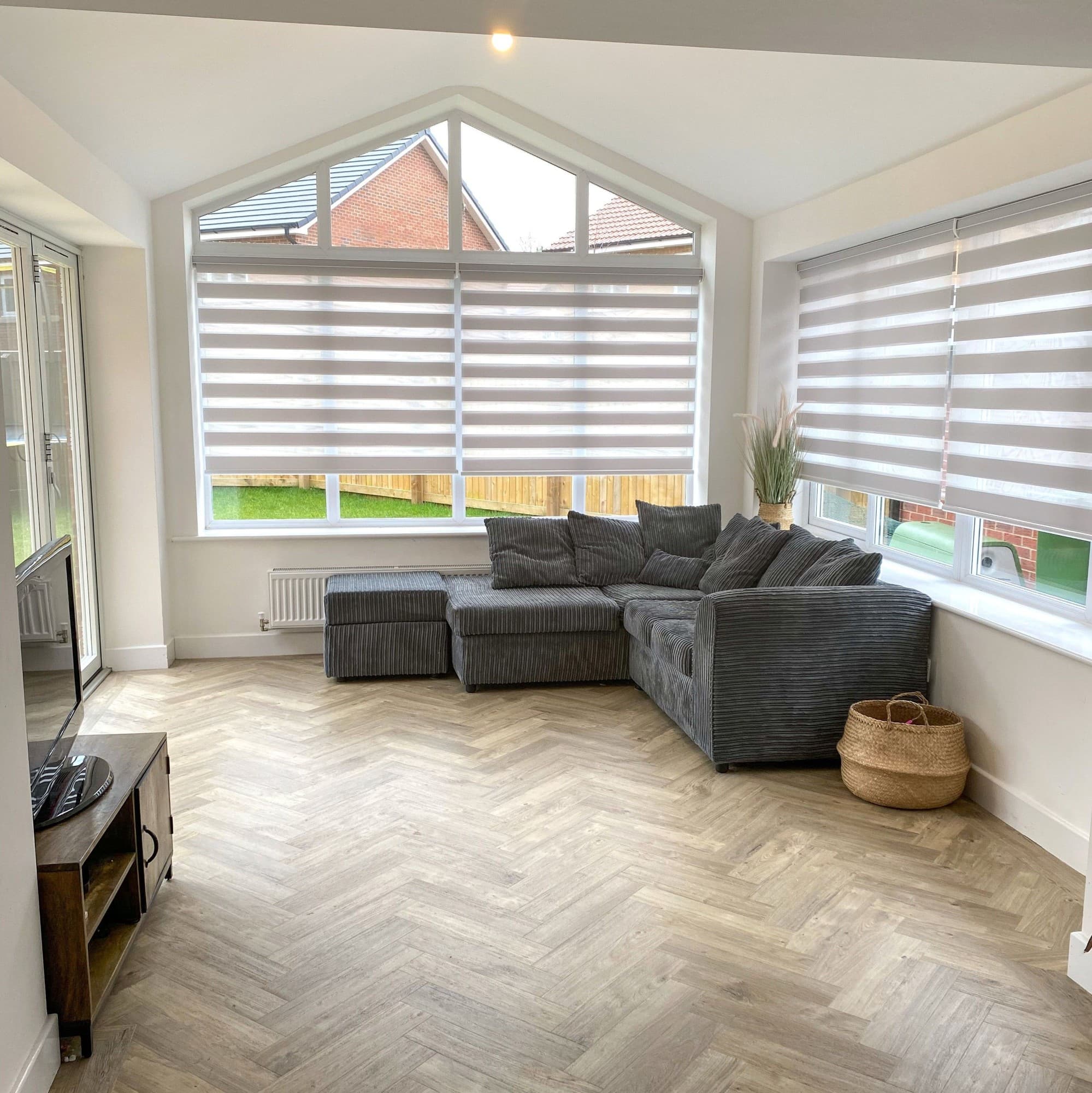 Zebra roller blinds covering triangular-topped bay and side windows, lowered and filtering daylight in a bright living room with a grey corner sofa, wood herringbone floor, overlooking a lawn.