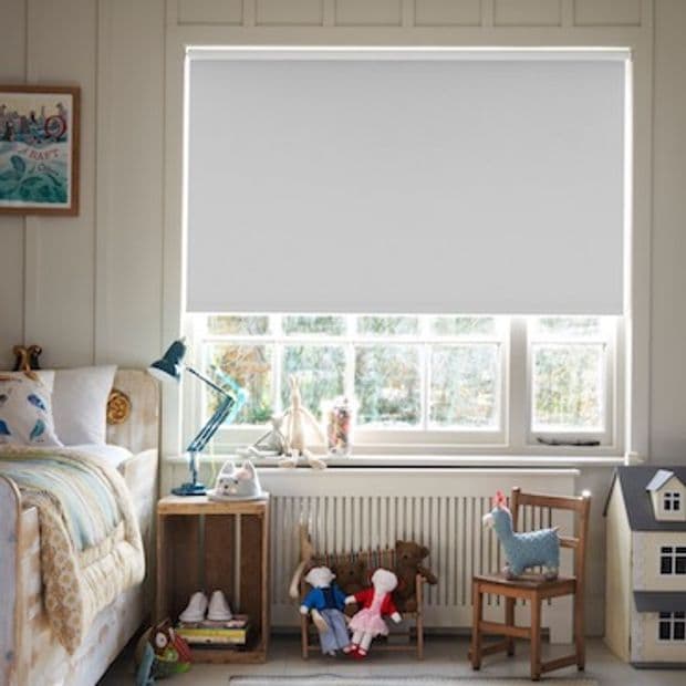 Roller blind (light grey, opaque) pulled down across upper window, diffusing daylight while lower panes remain visible. Childâ€™s bedroom with bed, lamp, toys and small garden view beyond.