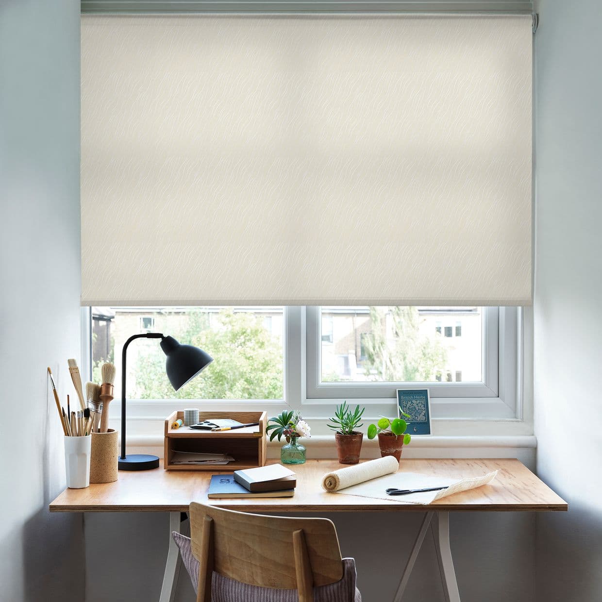 A beige patterned roller blind, lowered and filtering daylight; covering a double window above a wooden desk workspace with lamp, art brushes, notebooks, small potted plants and suburban view.