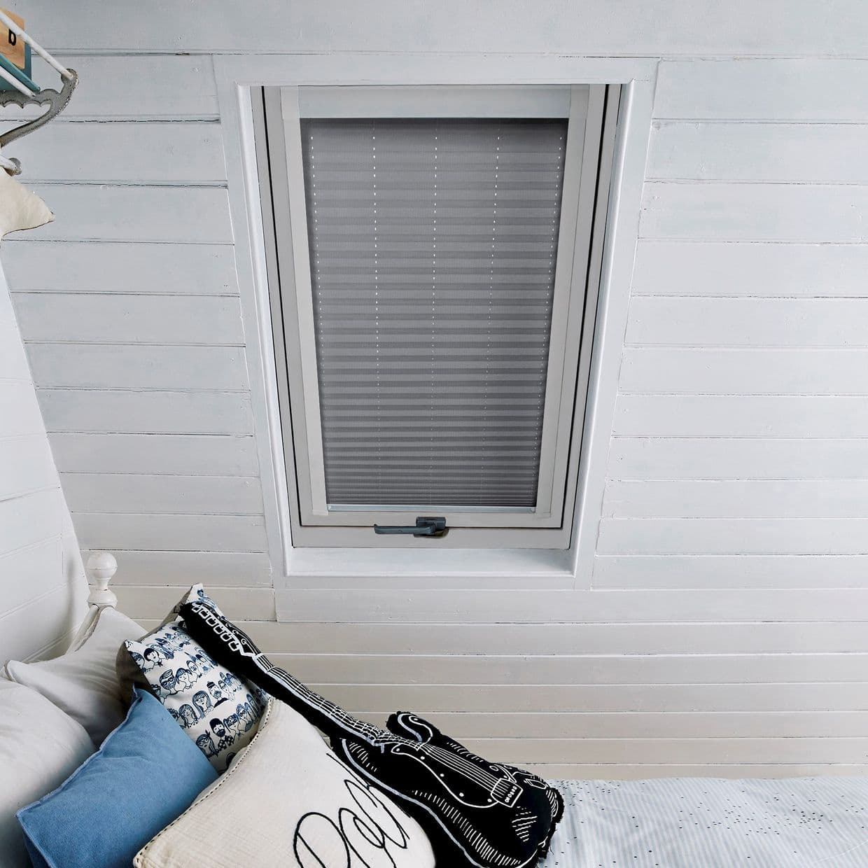 A gray pleated cellular blind pulled closed over a skylight, dimming light; set in a white-painted tongue-and-groove attic ceiling above a bed with patterned pillows.