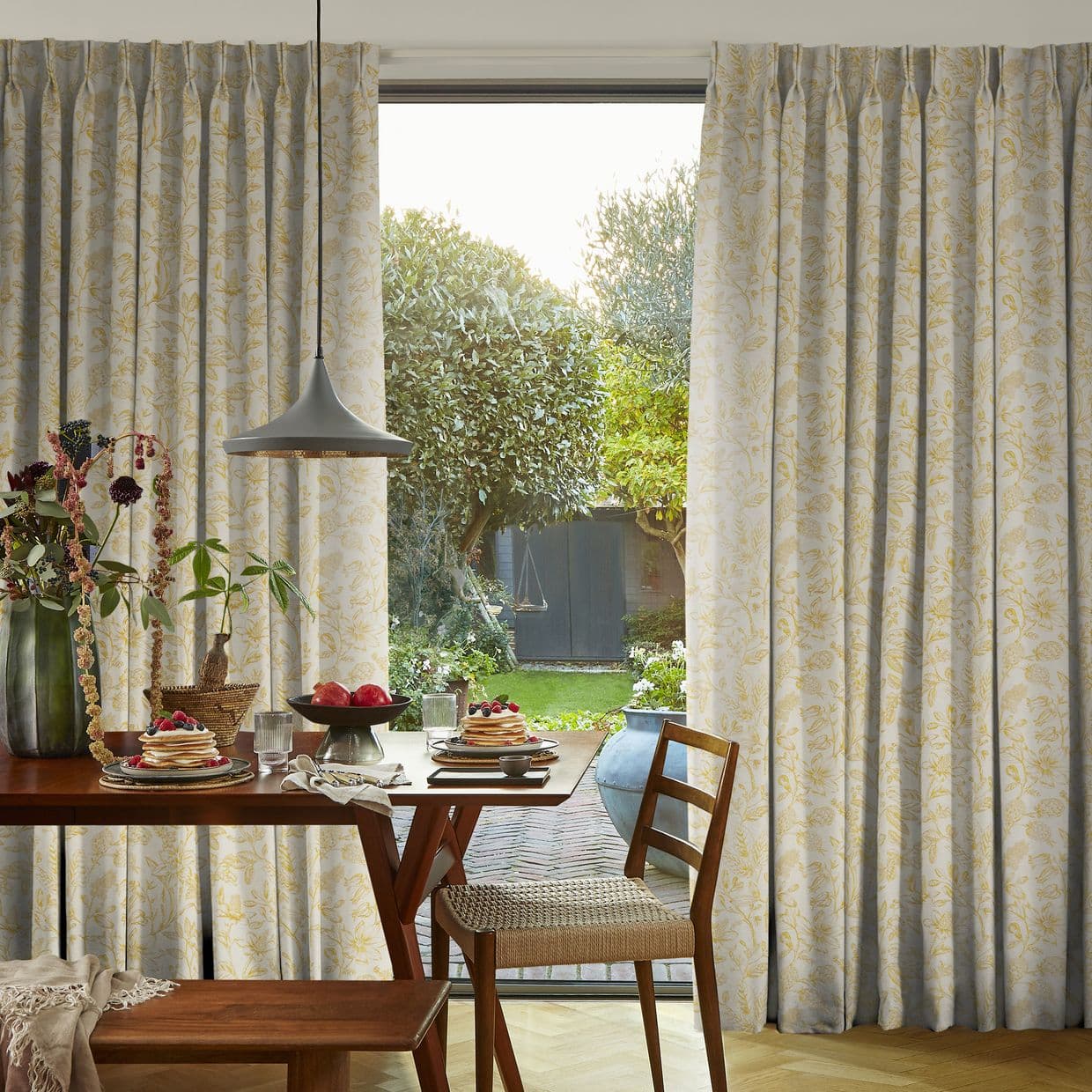 Full-length floral curtains in cream with pale yellow vines, framing and partially drawn aside at a sliding glass door, revealing a sunlit garden beyond; dining table set with breakfast inside.