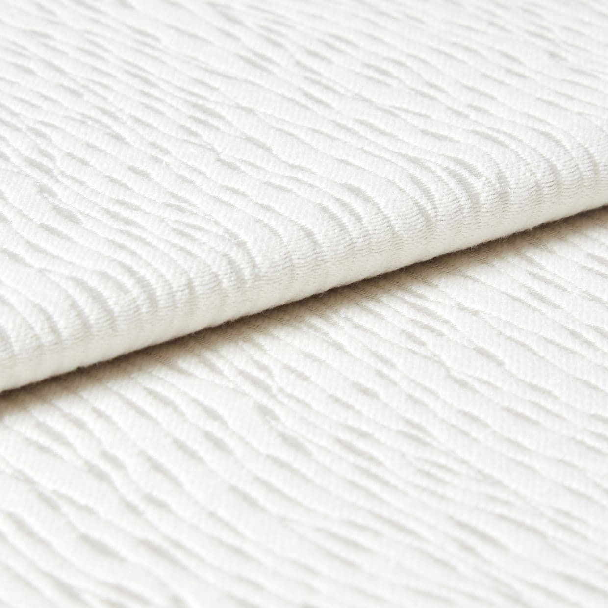 White wavy-ribbed fabric folded with an overlapping edge; fabric lies flat and layered. Close-up view under soft, diffuse light on a neutral surface, suggesting bedding or upholstery.