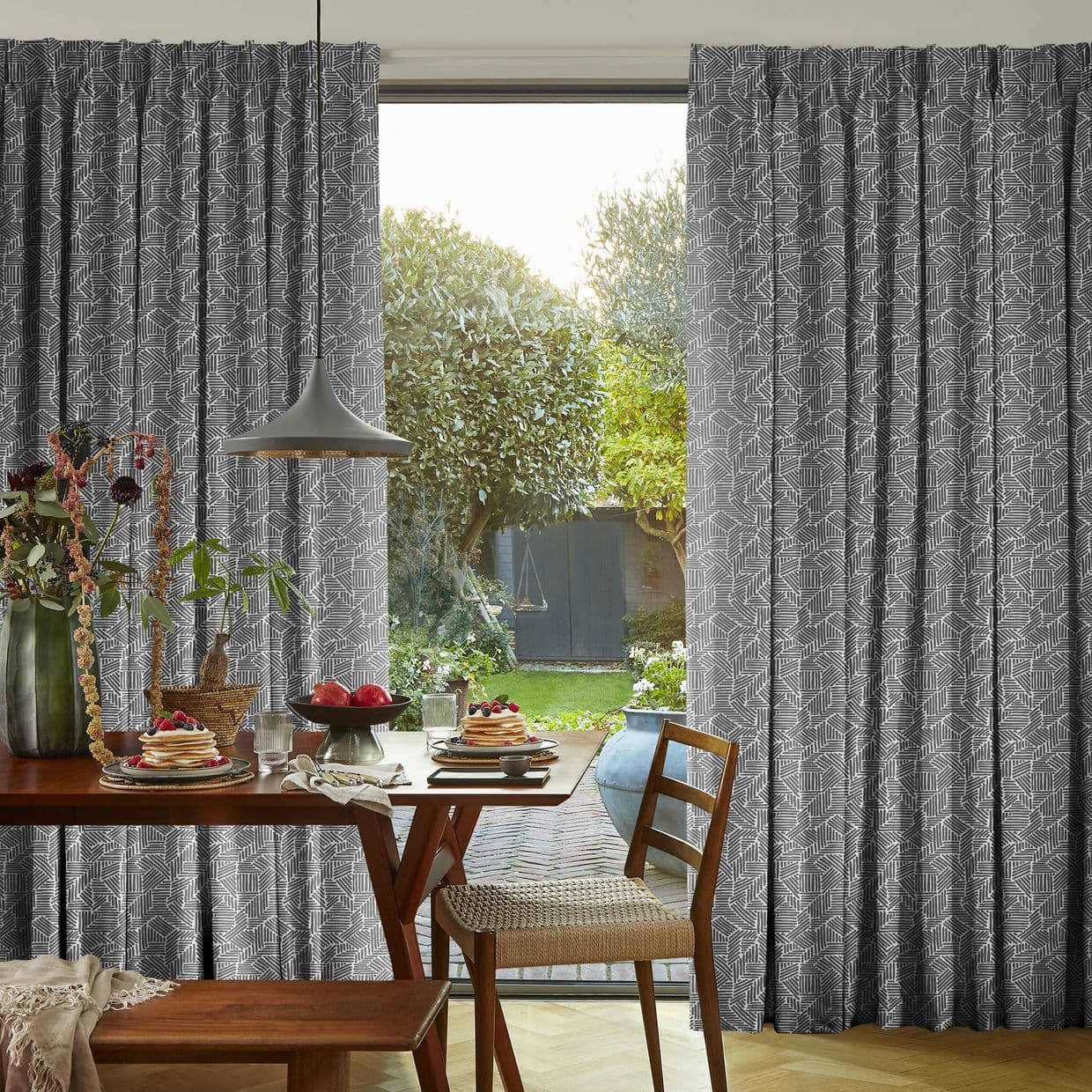 Patterned grey geometric curtains, pleated and drawn aside, framing and filtering sunlight; a wooden dining table set for breakfast with pendant lamp, opening onto a sunlit garden with trimmed trees.