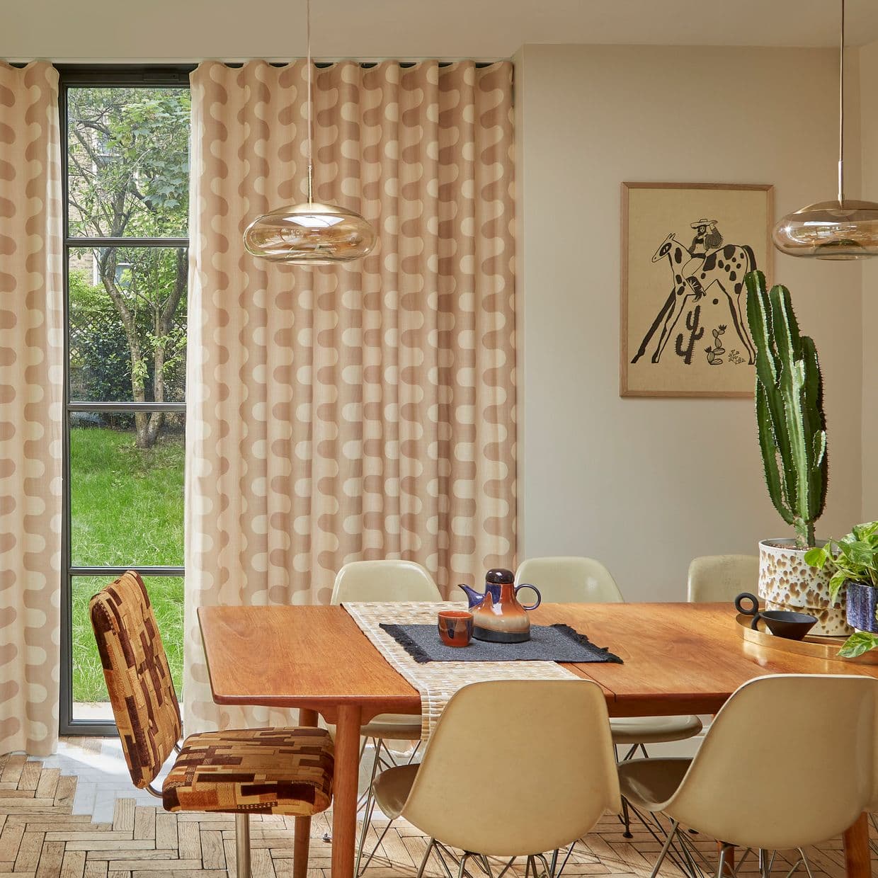 Beige grommet-top curtains with cream wavy pattern, mostly drawn and softly filtering daylight; dining room with wooden table, pendant lights, potted cactus, and garden visible through a narrow window.