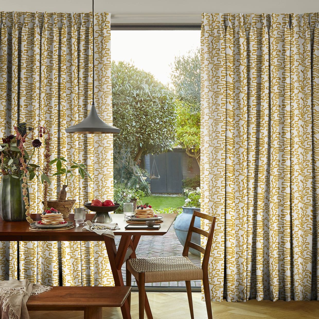 Patterned pleated curtains (white with mustard-yellow squiggles) hung, parted to frame a glass door; they filter daylight over a dining table set with pancakes, a pendant lamp and garden view.