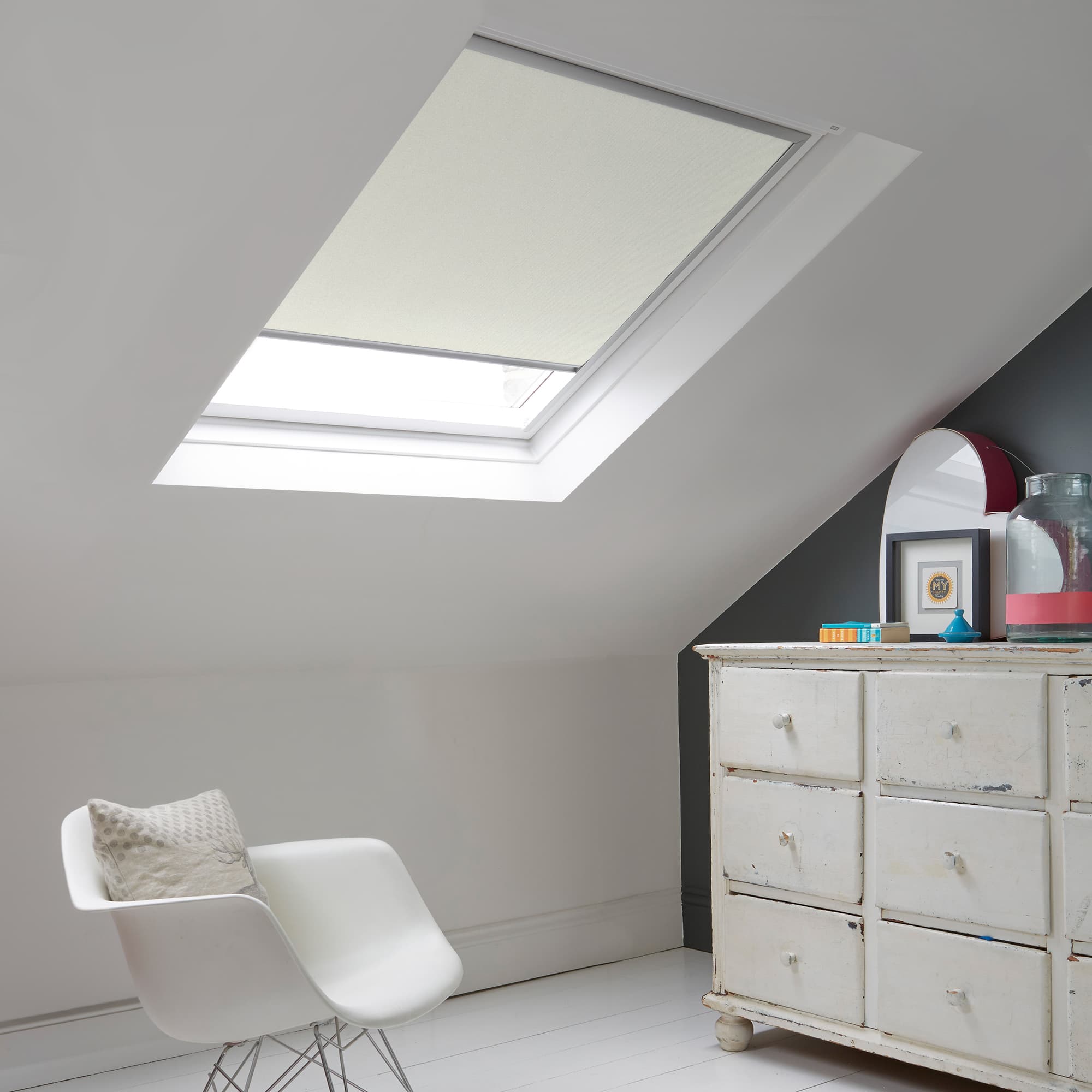 Room with white eaves wall, dark blue-grey wall to right with a shabby chic white drawer unit, a contemporary white chair, white painted floorboards and a skylight blind in pearl with a subtle glitter effect.