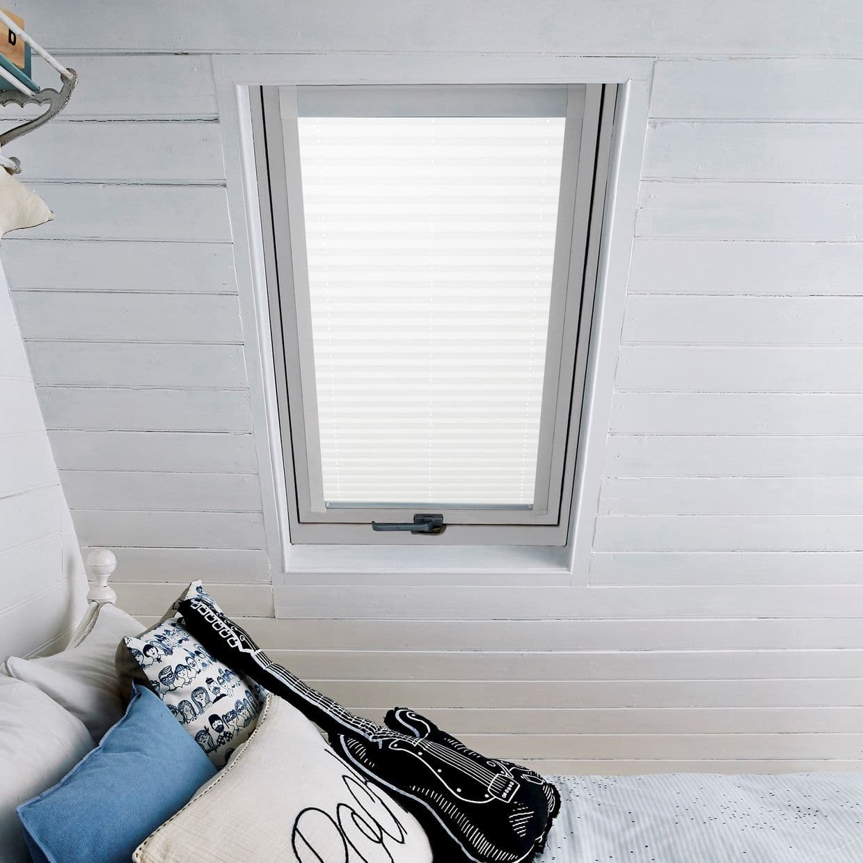 White pleated blind covering a skylight window, fully lowered and diffusing daylight; set within white-painted wooden sloped ceiling above a bed with blue and patterned pillows and a guitar-shaped cushion.