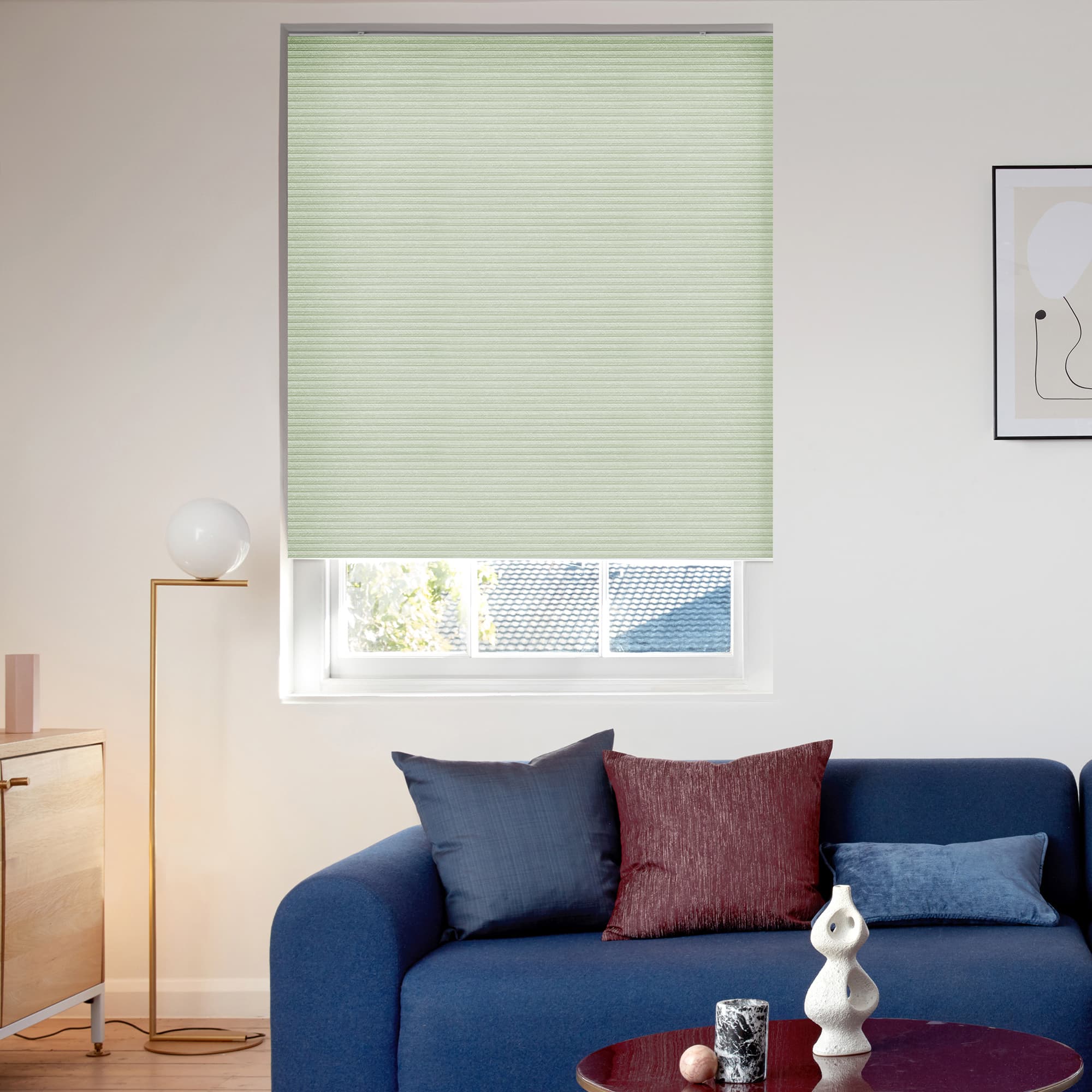 A pale green pleated roller blind, fully lowered with subtle horizontal ribs, filtering daylight; positioned over a window in a bright living room above a blue sofa with cushions.