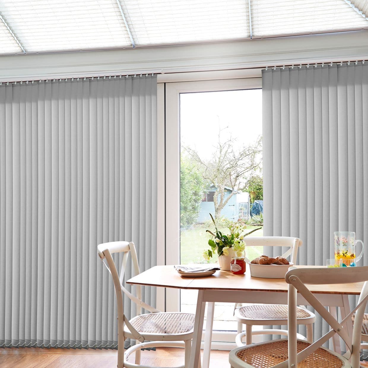 Light-gray vertical blinds on a ceiling-mounted track, slatted and mostly closed with a central gap exposing a patio door; filtering daylight into a dining area with table and garden view.
