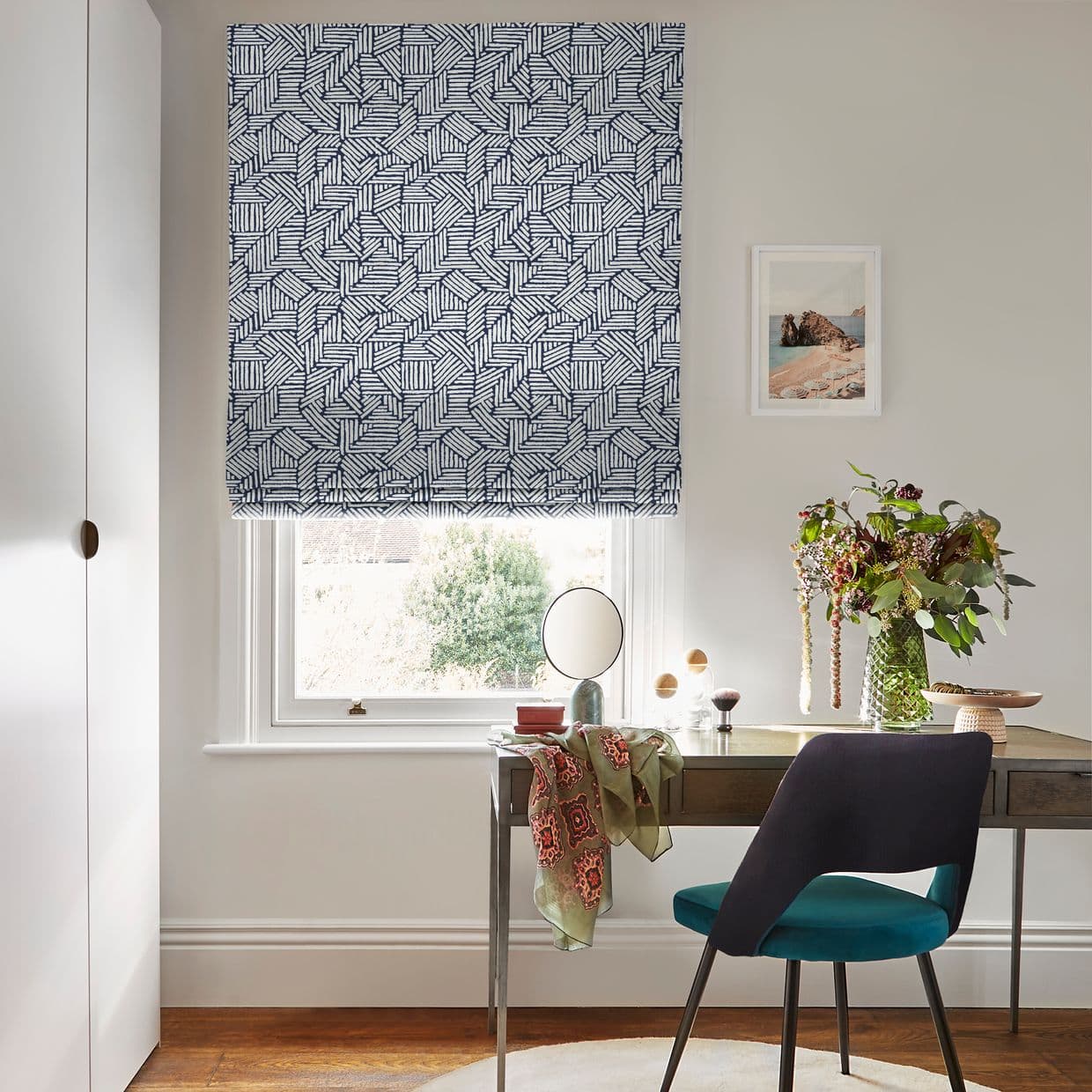 A navy-and-white geometric Roman blind, lowered and filtering soft daylight over a window; a desk beneath holds a mirror, vase of flowers, and a teal chair in a neutral office.