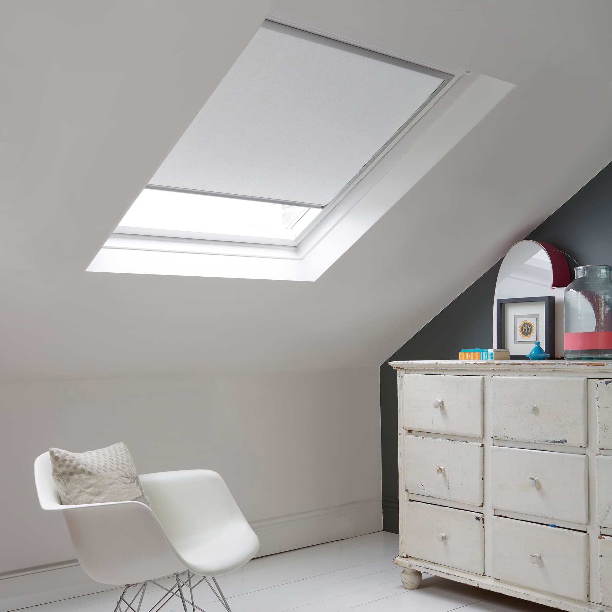 Room with white eaves wall, dark blue-grey wall to right with a shabby chic white drawer unit, a contemporary white chair, white painted floorboards and a skylight blind in pale mallow grey.