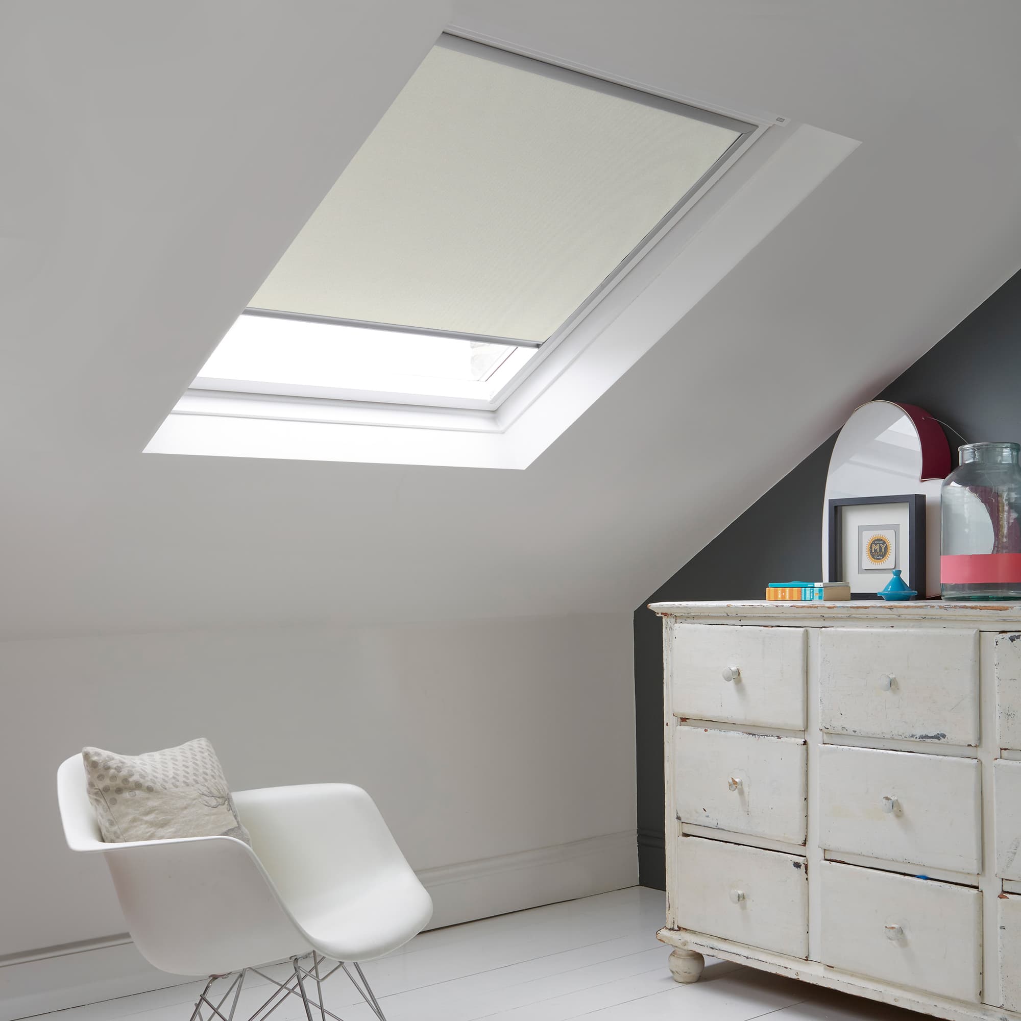 Room with white eaves wall, dark blue-grey wall to right with a shabby chic white drawer unit, a contemporary white chair, white painted floorboards and a skylight blind in off-white.