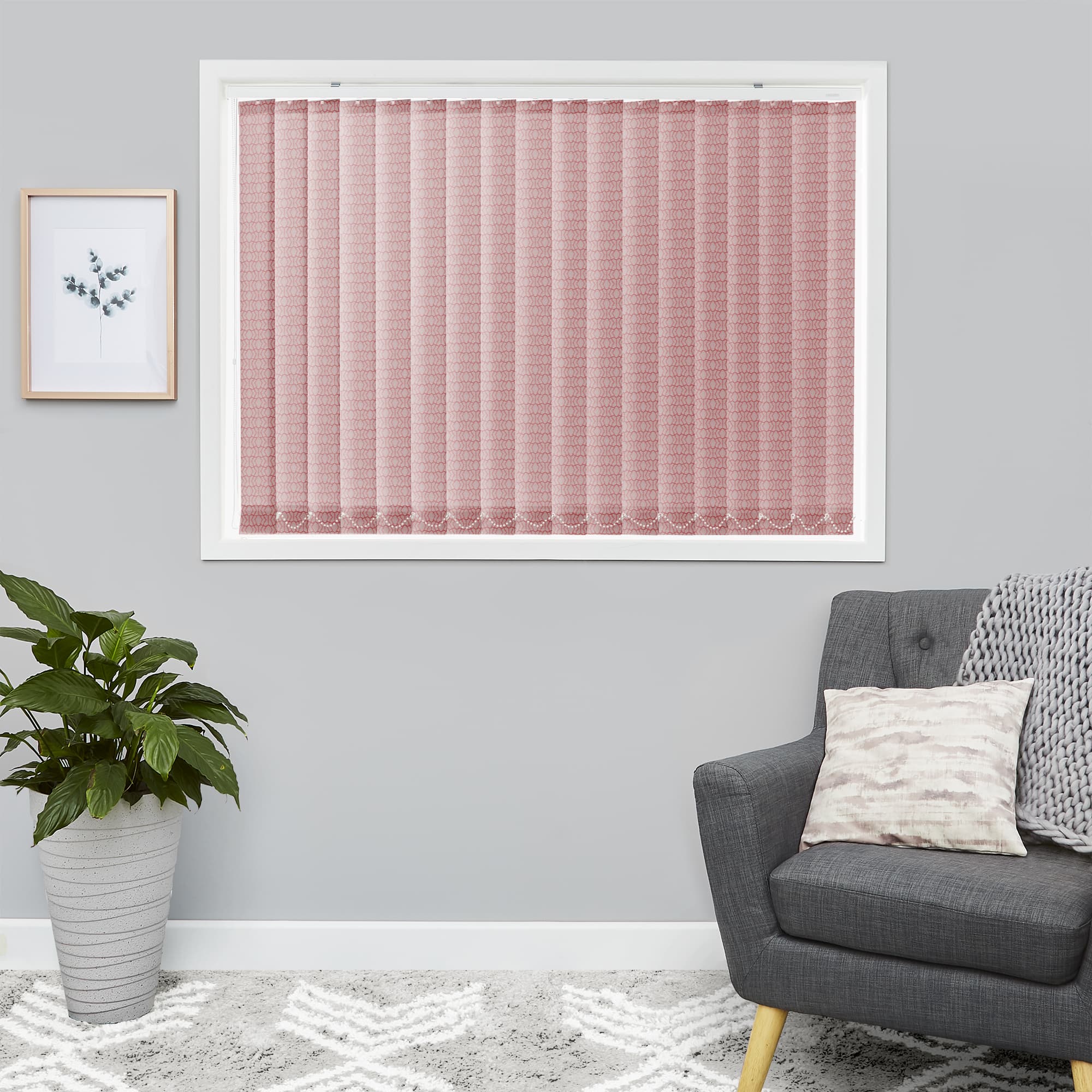 Pink patterned vertical blinds fully closed over a white-framed window, filtering soft daylight; in a gray-walled room corner with a gray armchair, knitted throw, potted plant and framed botanical print.