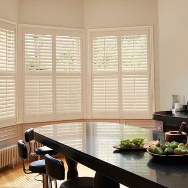 White plantation shutters covering a three-panel bay window, full-height slatted panels closed and diffusing soft daylight; kitchen/dining context with glossy black island, bar stools, fruit bowls, and warm hardwood floor.
