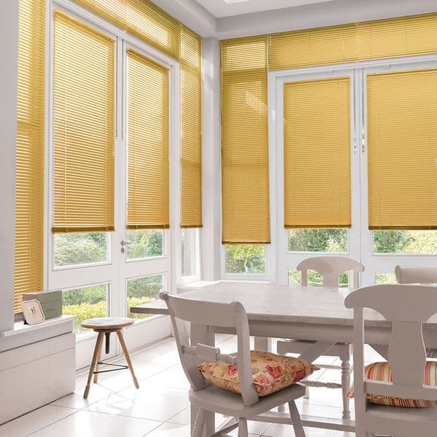 Yellow horizontal Venetian blinds covering tall bay windows, lowered and filtering warm sunlight; overlooking a green garden; bright dining nook with a white table, wooden chairs, and floral cushions.