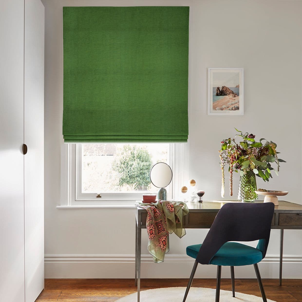 Green Roman blind â€” solid fabric with soft horizontal folds â€” is lowered, filtering daylight; a neat desk and teal chair sit beneath in a bright, minimalist home office with flowers.