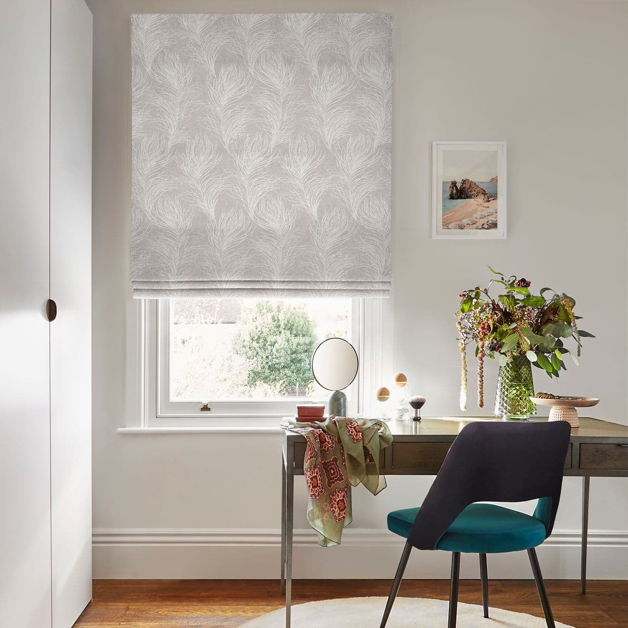 Pale-grey Roman blind with white feather motif lowered over the window, filtering daylight; desk with mirror, teal chair and vase of flowers sits against neutral walls beneath small seascape.
