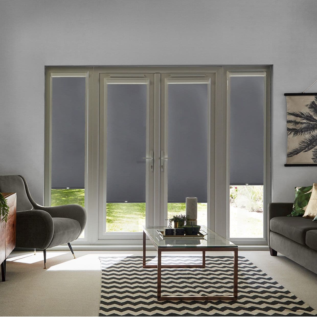 Dark grey roller blinds, four over French doors and sidelights, lowered to cover glass leaving small bottom gaps, filtering light in a living room with sofa and coffee table, lawn beyond.