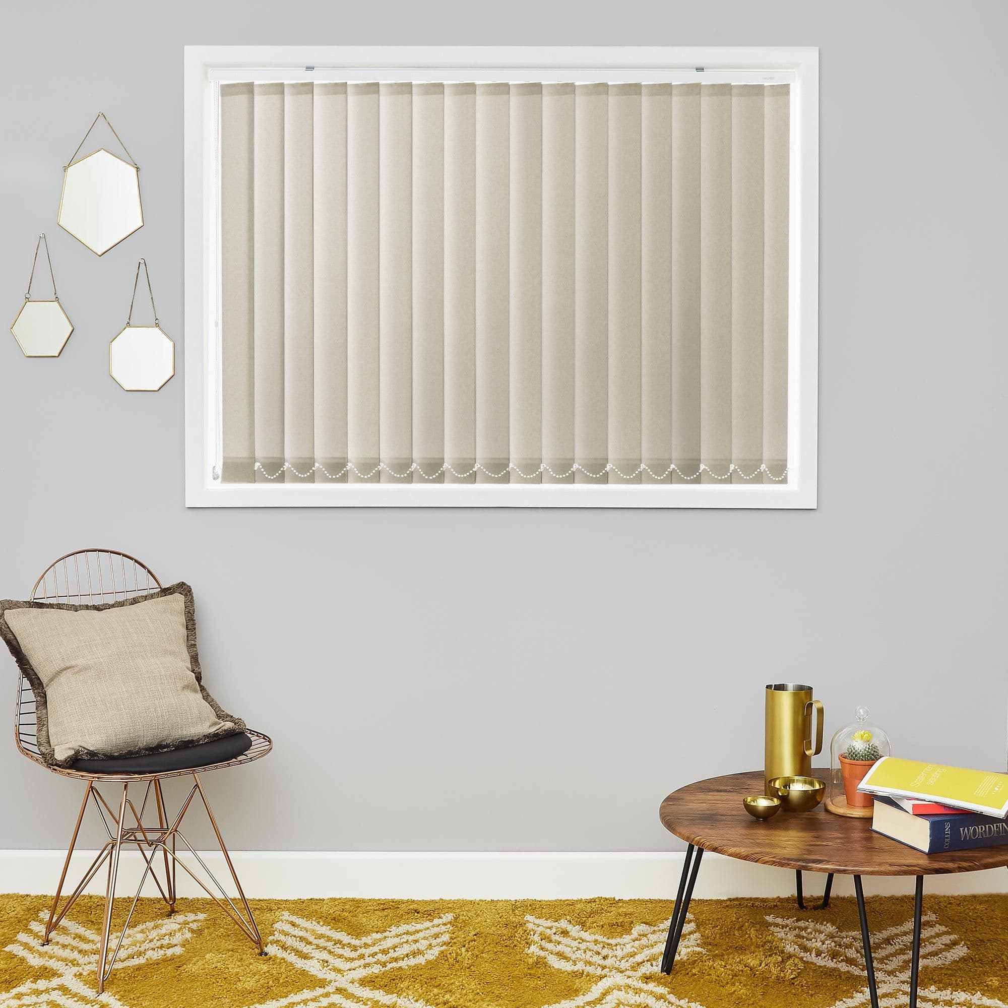 Beige vertical blinds covering a white-framed window, closed and lightly diffusing daylight; set in a grey-walled living space with a wire chair, cushion, wooden coffee table, and mustard patterned rug.