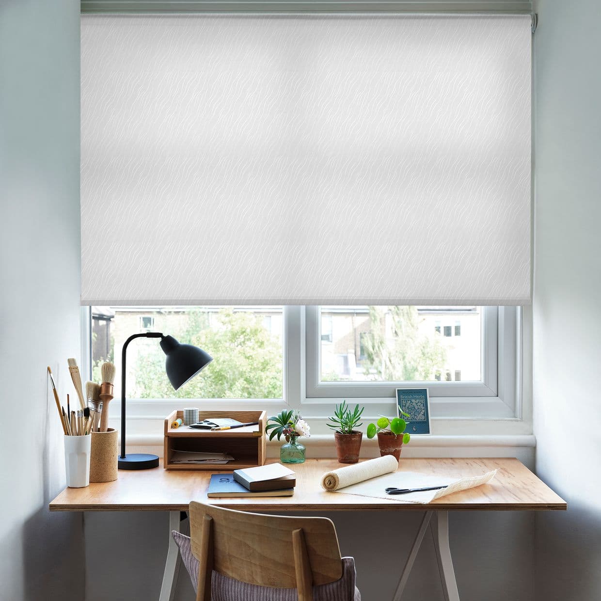 A white patterned roller blind, lowered across a double window, filtering daylight; above a wooden desk with lamp, brushes, notepads and small potted plants in a bright home workspace.