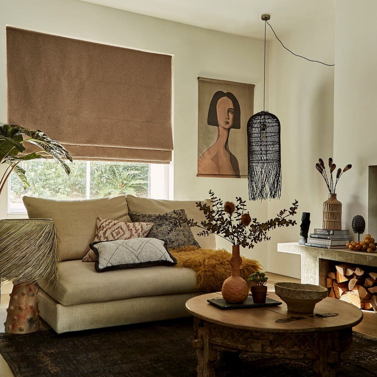 Brown Roman blind lowered over a window, filtering daylight; in a warm neutral-toned living room with a beige sofa, patterned cushions, wooden coffee table, pendant lamp and wall art.