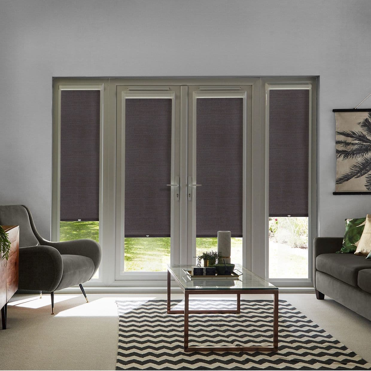 Dark fabric roller blinds cover four-panel French doors, partially lowered and filtering daylight; in a living room with a grey armchair, sofa, glass coffee table on a zigzag rug, garden view.