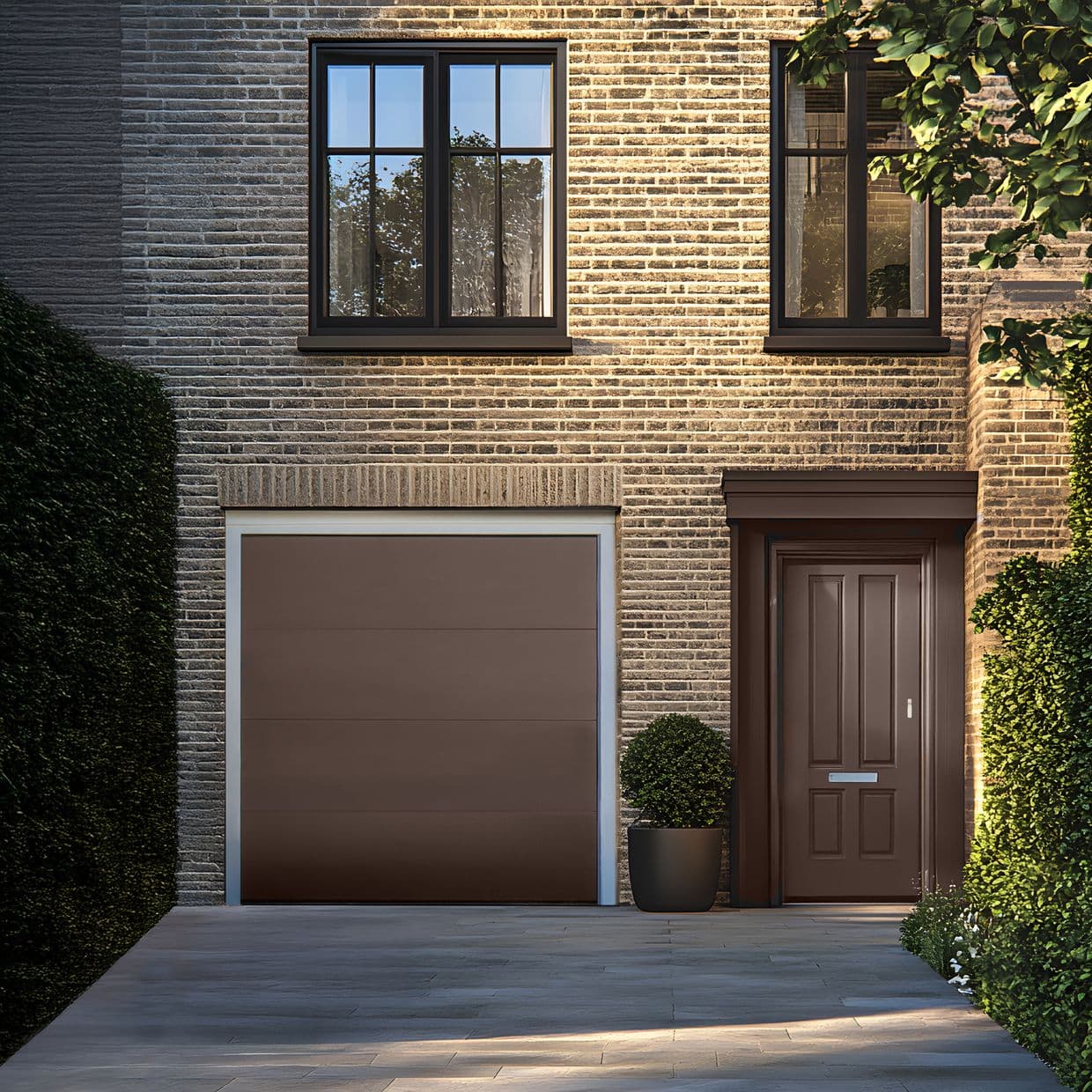 Sheer curtains in the upper-left black-framed window, partly drawn and filtering daylight; set on a brick townhouse facade with matching brown garage and front door, potted shrub and shaded driveway.