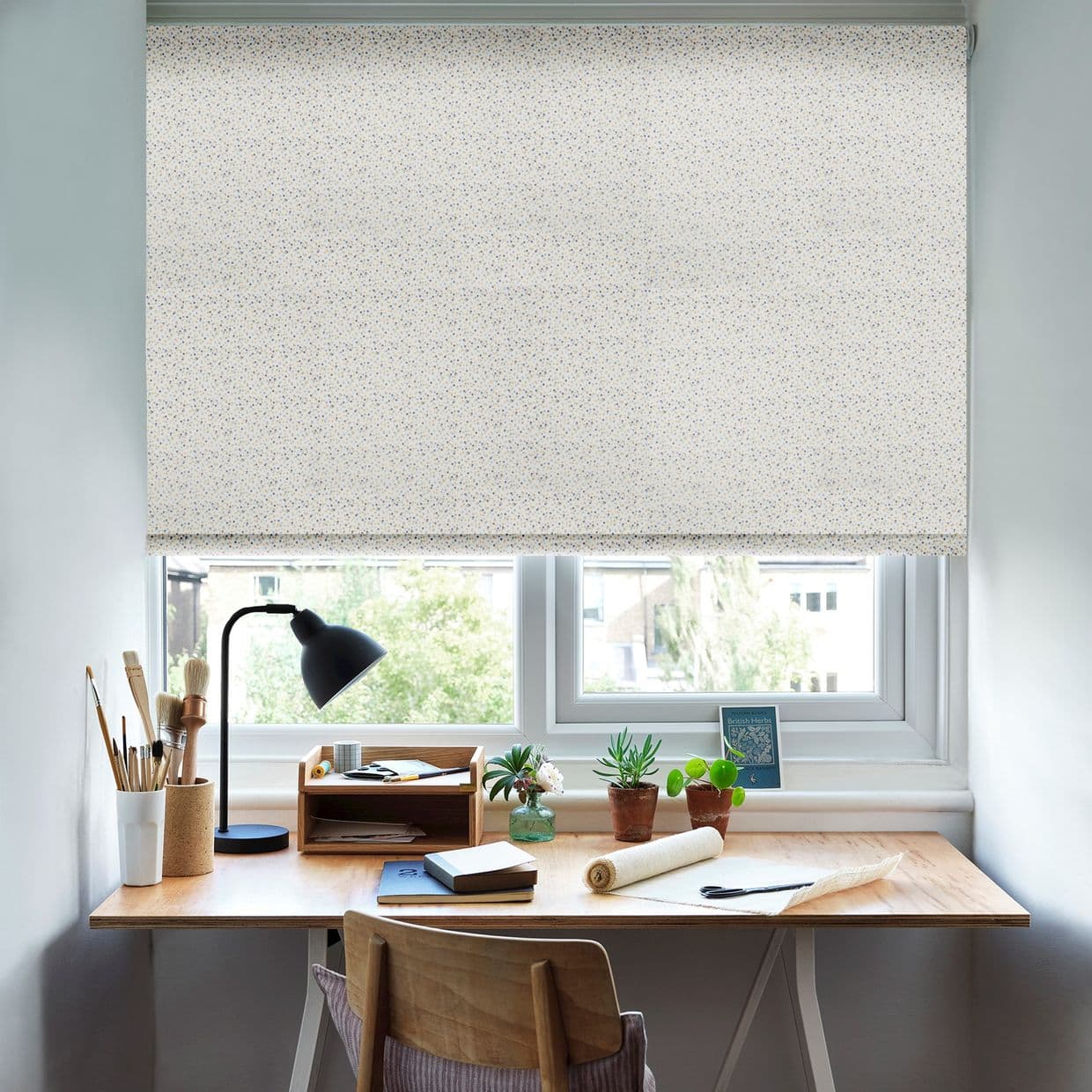 A patterned roller blind, fully lowered with tiny blue-yellow speckles, diffusing daylight and blocking the view; below it sits a wooden desk with a lamp, potted plants, and stationery in a bright home office.