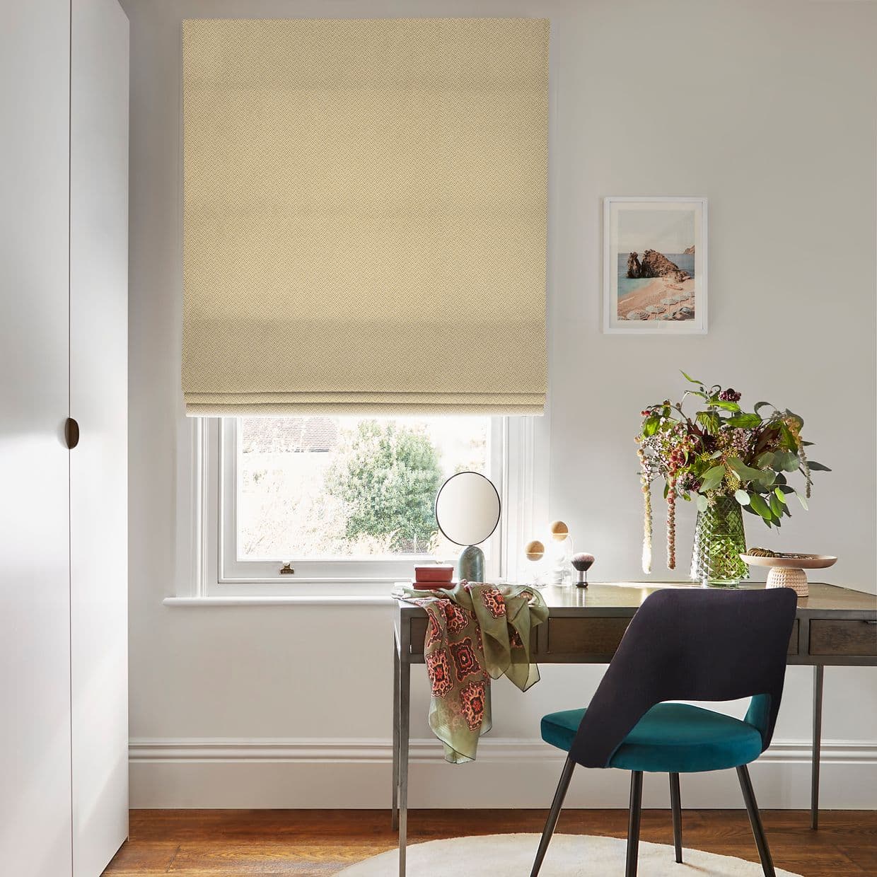 Beige patterned Roman blind pulled down over a white-framed window, softly filtering daylight; below, a wooden desk with mirror, vase of flowers and teal chair in a bright, neutral home office.