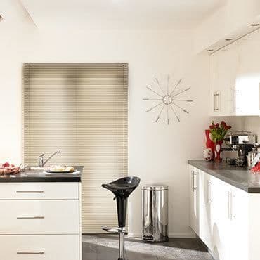 Beige horizontal Venetian blinds, fully closed, diffusing soft daylight through kitchen window; white kitchen with grey countertops, chrome trash bin, black barstool, wall clock and red flowers on counter.