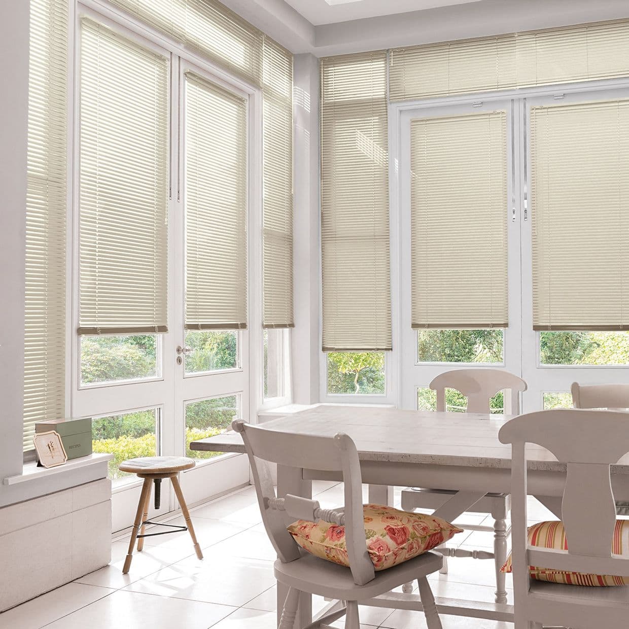 Beige horizontal Venetian blinds covering tall French windows and doors, lowered and filtering soft daylight; bright, sunlit dining nook with white wooden table, chairs, floral cushions, and garden view.