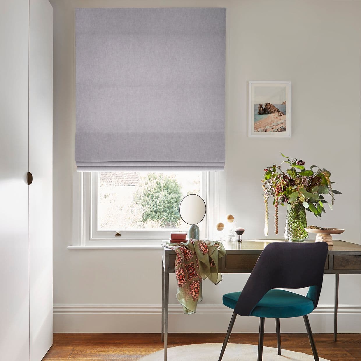 A light grey roman blind lowered over a sash window, soft-folded fabric gently filtering daylight; below, a desk with mirror, vase of flowers and teal-upholstered chair in a home office.