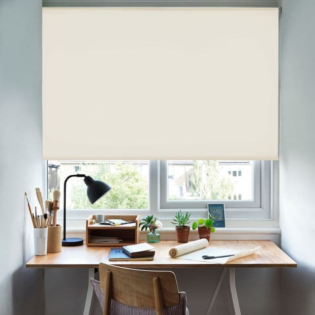 Beige roller blind lowered over a double window, smooth fabric blocking upper light and diffusing daylight; home study desk beneath with lamp, wooden chair, potted plants, notebooks and art supplies.