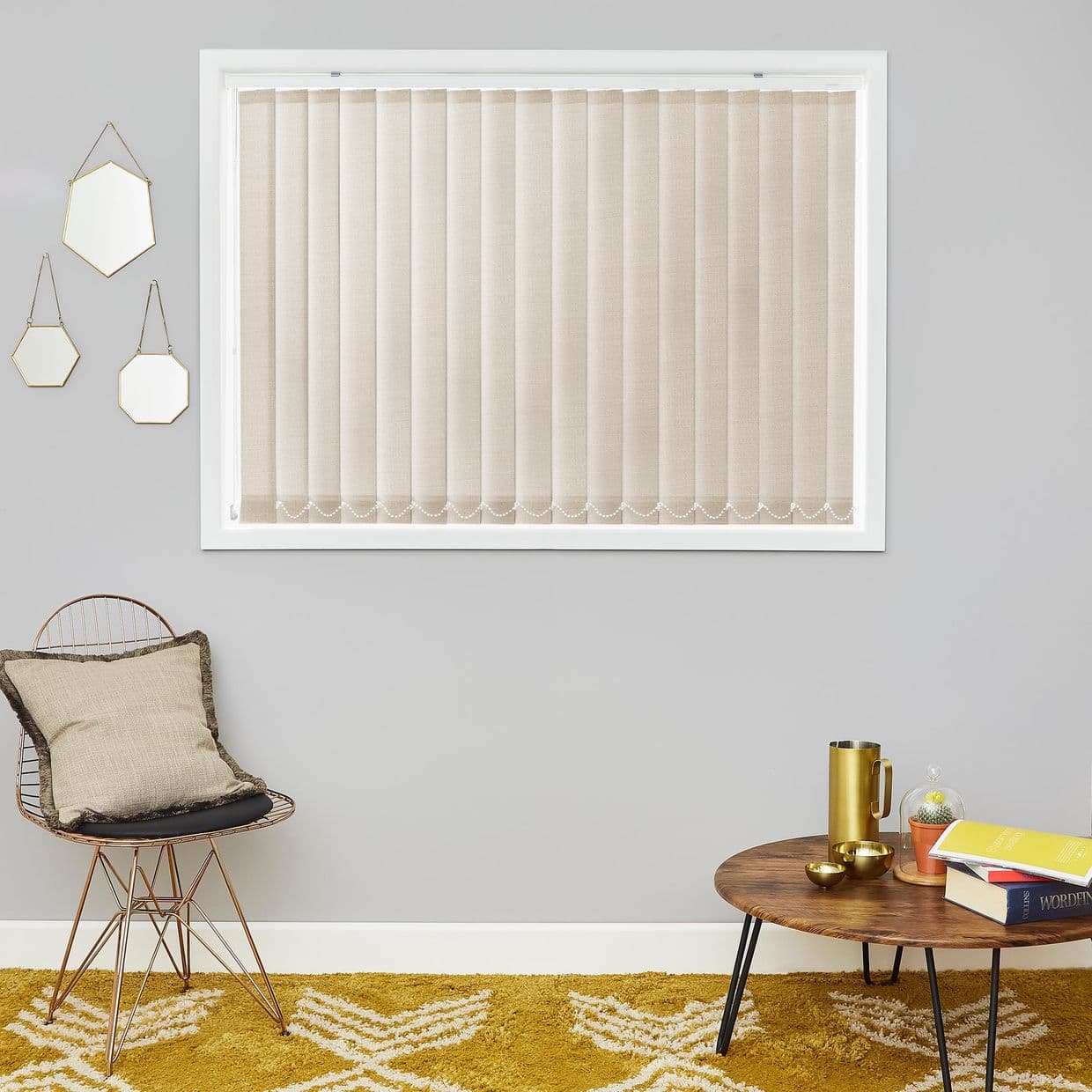 Beige vertical blinds fully closed across a white-framed window, evenly spaced slats with chain weights at bottoms, filtering daylight into minimalist living room with grey walls, chair and coffee table.
