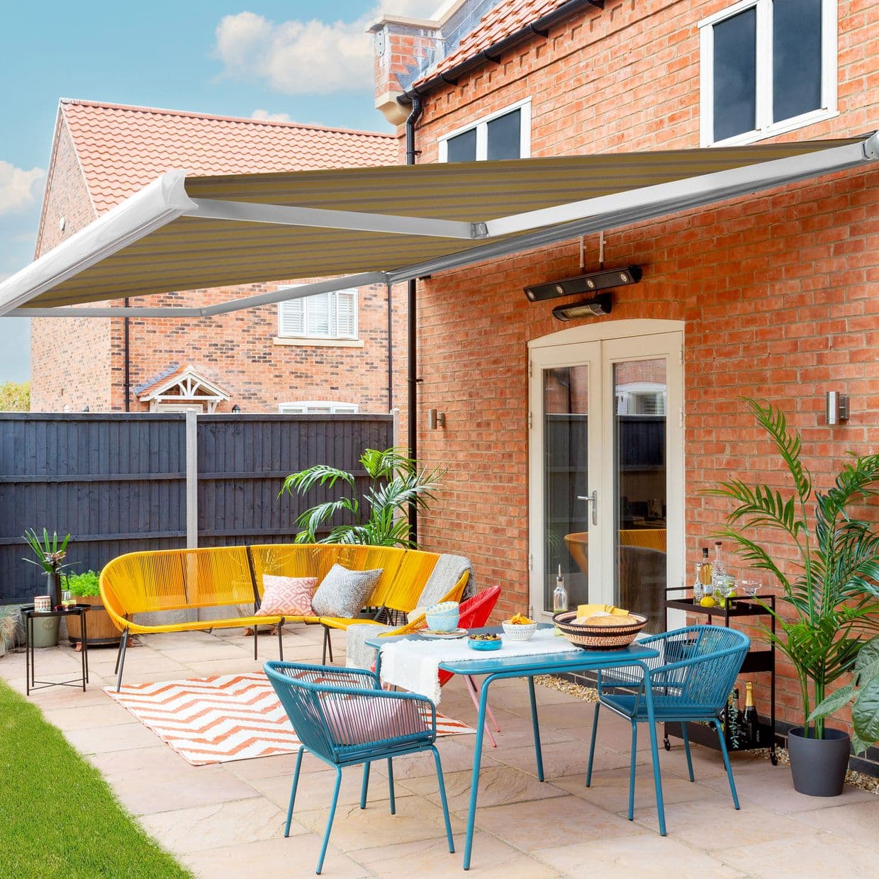 Striped retractable awning, mounted above French doors, extended with metal arms providing shade over a sunny brick-house patio with colorful furniture, rug and potted plants.