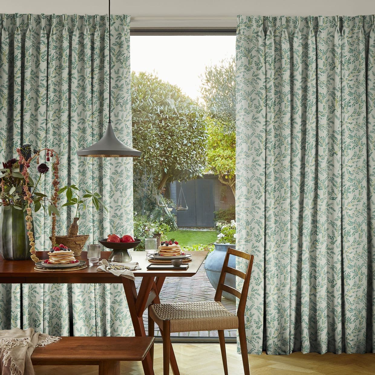 Patterned pleated curtains with green leaf print, parted to frame a glass sliding door and let warm sunlight in, revealing a breakfast table and a lush backyard garden.