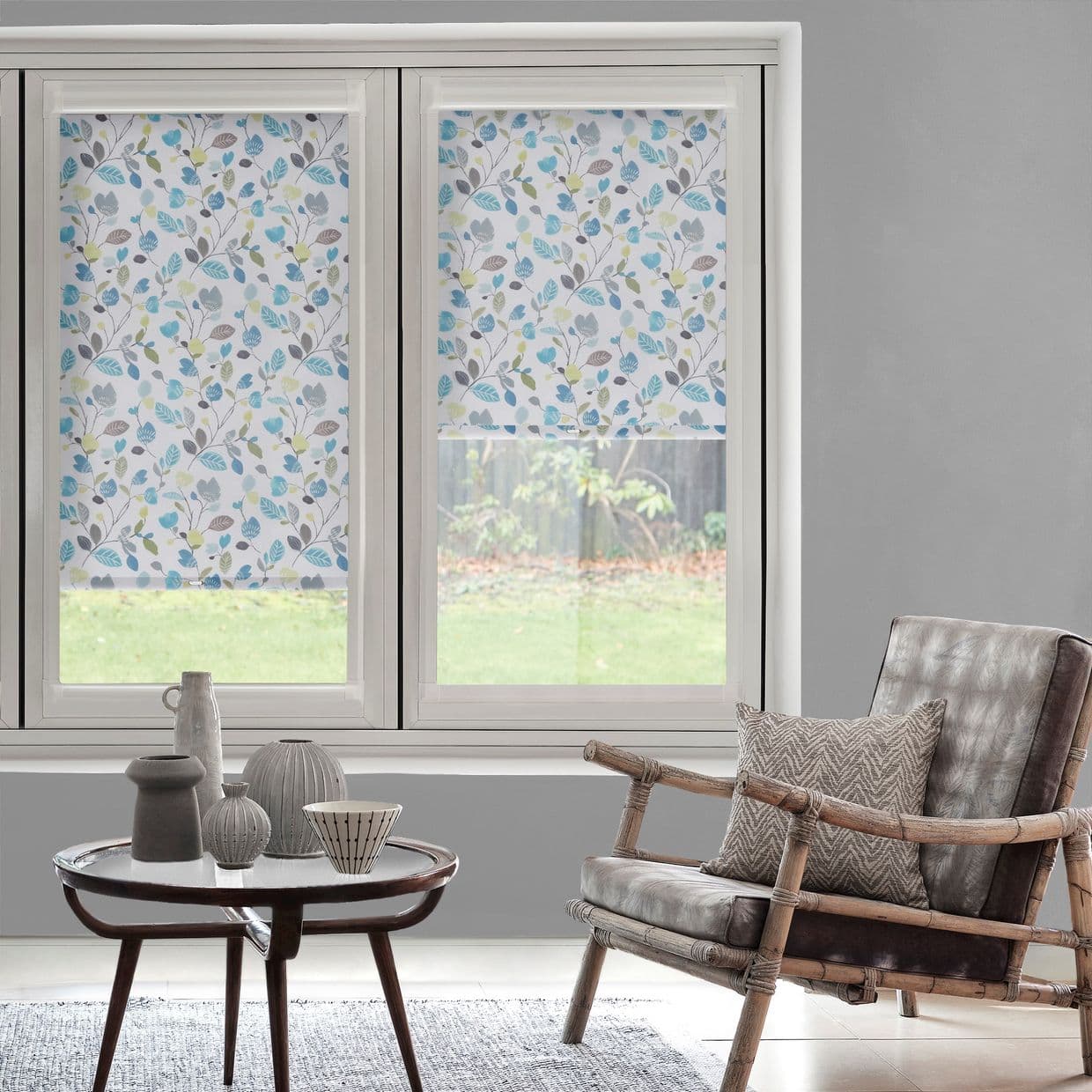 Floral leaf-pattern roller blinds covering twin windowsâ€”one fully lowered, one partiallyâ€”filtering daylight; bright living room with gray walls, rattan armchair, round wooden table with ceramics, backyard lawn visible.