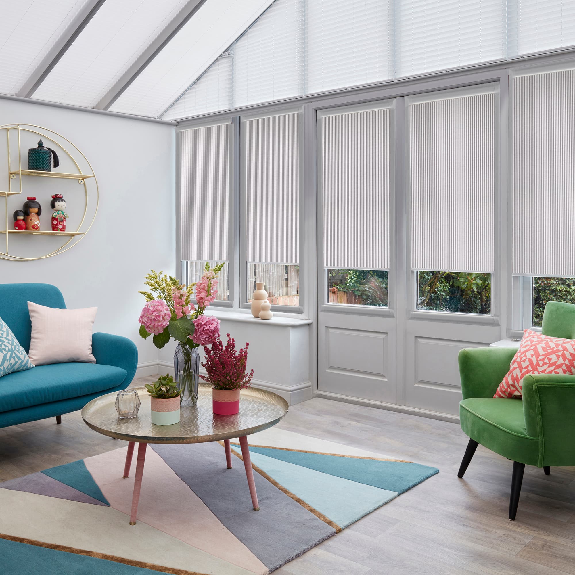 White pleated cellular blinds lowered across tall conservatory windows and door, filtering soft daylight; living room with teal sofa, green armchair, floral centerpiece, geometric rug, garden view.