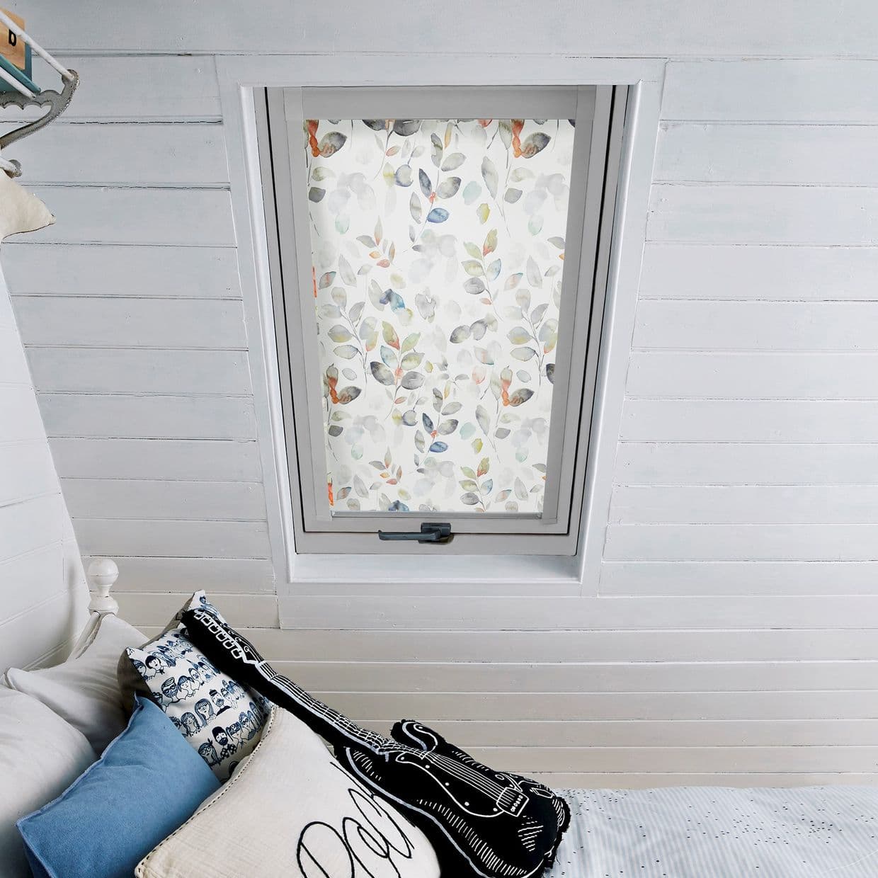 Floral roller blind with watercolor leaf pattern, fully lowered, diffusing daylight across a white-painted attic bedroom; slanted shiplap walls, bed with cushions and guitar-shaped pillow below.