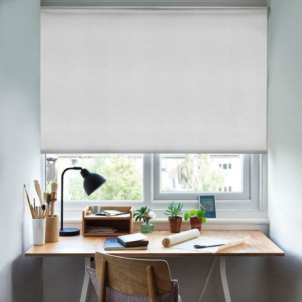 White roller blind lowered nearly to the sill, slightly translucent and smooth, filtering bright daylight; positioned over a double window above a wooden desk workspace with lamp, potted succulents, and stationery.